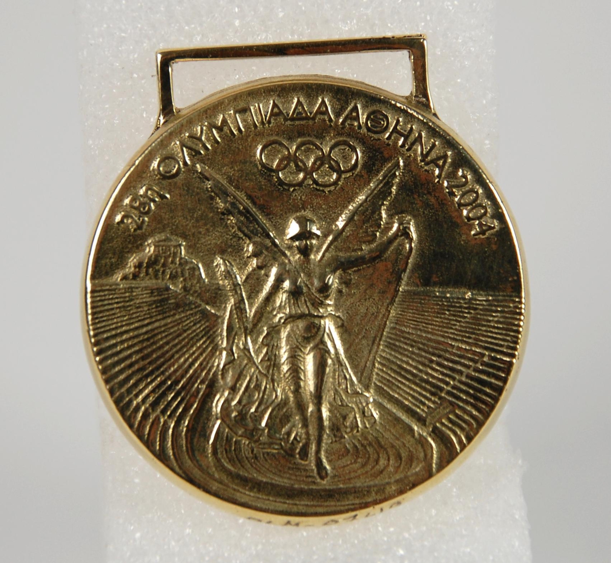 Gullmedalje med motiv av den greske seiersgudinnen Nike som flyr over en stadion. Medaljen har gresk innskrift.