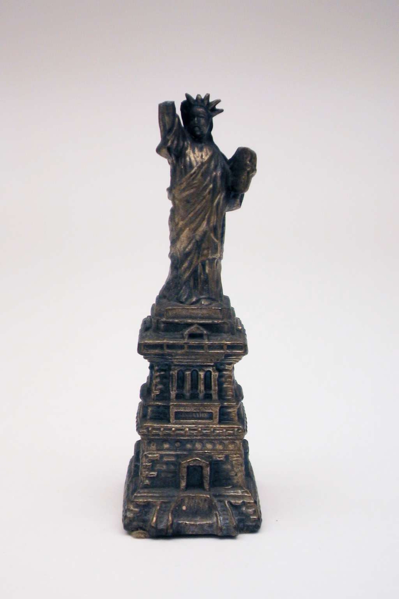 Den amerikanske frihetsstatuen i miniatyr. Den er laget i tinn som er bronsert. Den ene armen er defekt.