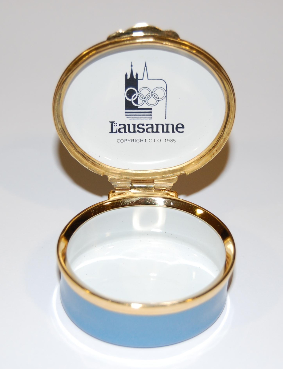 Flerfarget boks av metall med motiv av de olympiske ringene og Lausanne på lokket. Boksen ligger i en hvit eske av papp.