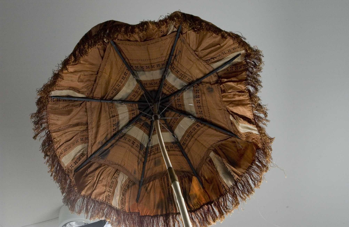 Liten brunmønstret parasoll med frynser langs kanten. Metall- og trespiler. Stangen er leddet og låses med en messingholk, malt hvit. Den nederst delen av stangen ser ut som bambus. Håndtak mangler. 