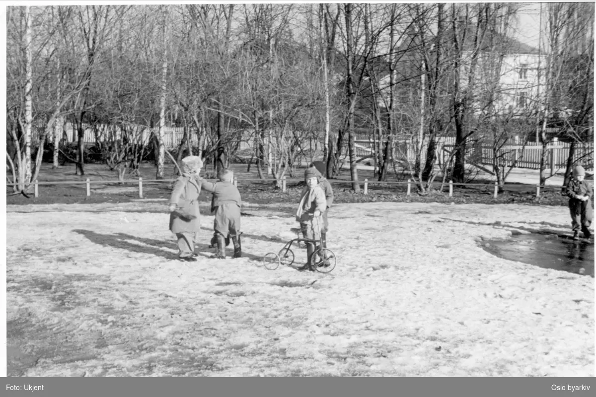 Vinter i Frognerparken. Lekende barn med en trehjulsykkel på skarasnø i enden av lekeplassen. Hvitt tre-stakittgjerde langs Kirkeveien.
