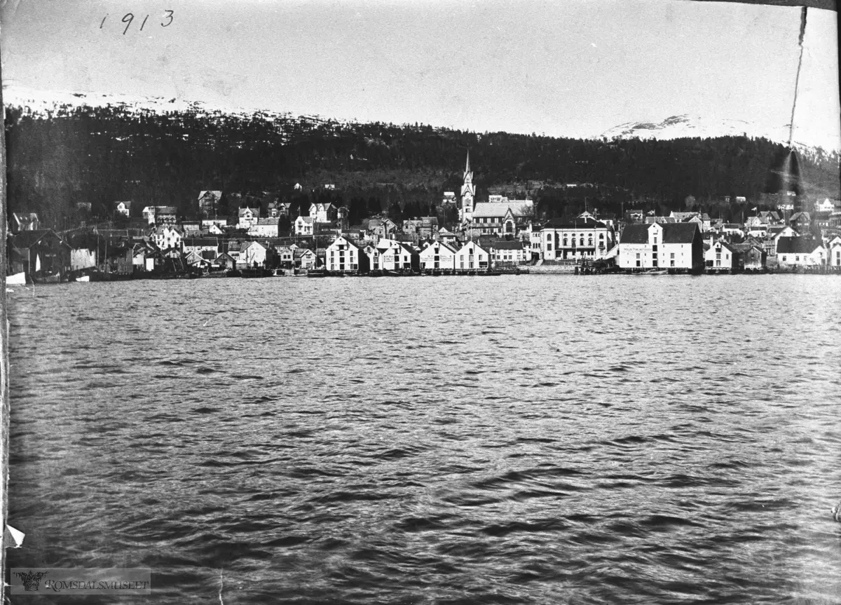 Molde by sett fra sør., Molde sett fra fjorden med kirken. Det står 1913 på bildet, men bildet er tatt i mars eller senere 1914. Vi ser to nedbrente brygger. Disse bryggene brant siste gang i mars 1914.