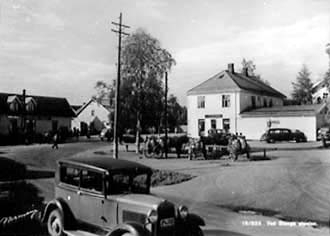 Postkort, Stange sentrum, Stangebyen, fra venstre: Magnus Godagers forretning i Storgata, og O, Nordsveens butikk til høyre, hestebinding på meieritorget, hester med stuttkjerrer kjører mjølk, en bil: Ford i forgrunnen med registreringsnummer D-4358, i heftet Hedmarks Bileiere 1938 er bilen registrert på Ole Wie,