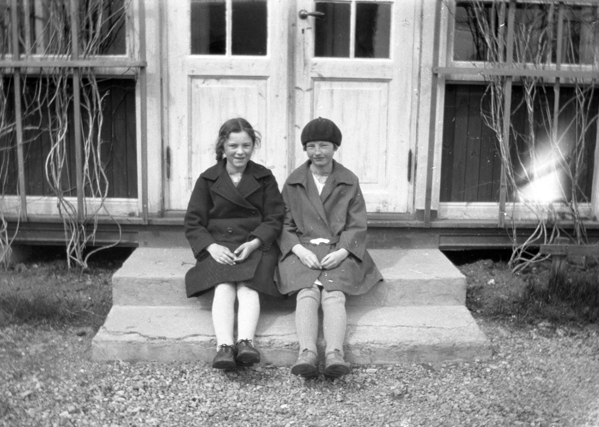 To jenter i kåper sitter på ei trapp.
Bildet er tatt på Nordtun gnr. 31, bnr. 178 i Elverum, nåværende gateadresse Strandbygdvegen 105. Jentene er fra venstre: Frida Skibness f. 1916 og Margit Syljeset f. 1915. Frida vokste opp på Nordtun.