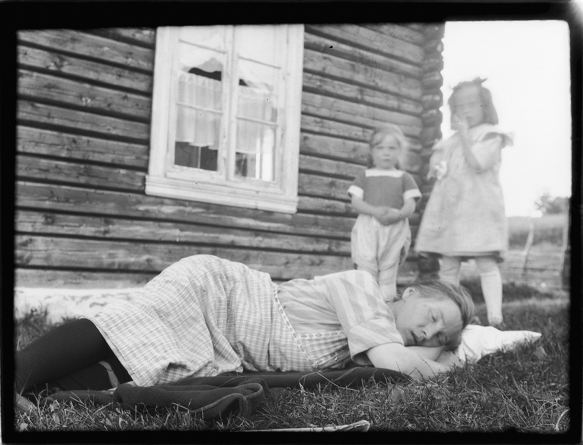 Ukjent kvinne sover i gresset, to barn i bakgrunnen. 
