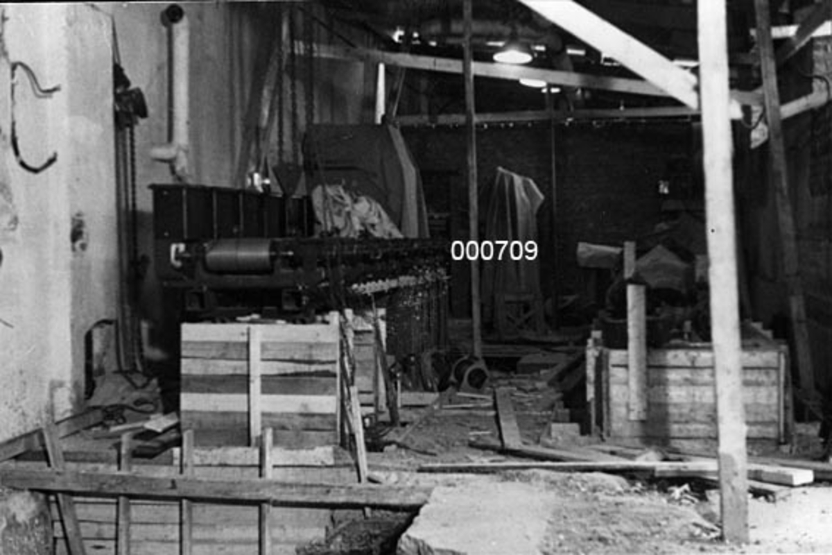 Interiør fra A/S Buskerud Papirfabrikk på Åssida i Drammen (den gang i Lier kommune).  I 1937 ble papirmaskinen ved denne fabrikken fullstendig ombygd, særlig for å kunne kjøre maskinen med større driftshastigheter, slik at en kunne oppnå større produksjonsvolumer.  I den forbindelse la bedriften om fra dampdrift til elektrisk drift.  Fotografiet er tatt i transmisjonsrommet, der kraft fra bedriftens dampmaskin var blitt overført fra en hovedaksling via diverste balatareimer til ymse maskindeler.  Da dette fotografiet ble tatt var rommet under ombygging, og mange av de gamle transmisjonene (akslinger med reimskiver) var borte.  Noe later imidlertid til å stå igjen langs veggen til venstre.  I forgrunnen later det til at noe er forskalet i en nedsenkning i golvet.  Taket støttes av stolper.  Dette er antakelig et midlertidig fenomen som var ledd i sikringstiltak i forbindelse med ombygginga. 