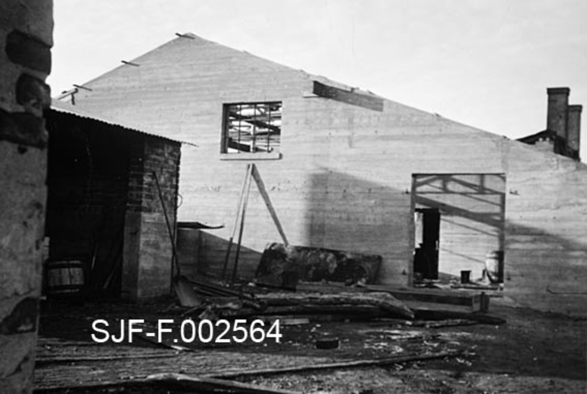 Byggearbeid ved fabrikkanlegget til Schwencke & Co's Eftf. på Børsholmen i Asker.  Fra 1924 drev dette firmaet framstilling av bek og andre tjærebaserte produkter på holmen. Virksomheten var ildsfarlig, og det oppsto flere branner.  Dette fotografiet er tatt i forbindelse med gjenoppbyggingsarbeider etter en brann. Den 9. februar 1938 kokte en av bekkokerne over og den varme massen rant nedover mot lavereliggende lagerskur. Der antente den lagerbygningen og beholdningen. Fotografiet er tatt mot gavlen på den lave, men brede nye kokeribygningen, som hadde støpte betongvegger. Taket var ennå ikke på plass, og ikke inngangsportene heller.  Til venstre i bildet ser vi et tilstøtende skur i teglmur. I forgrunnen ser vi skinnegangen til trallebanen, som ble brukt til å frakte gods mellom kaia nede ved stranda og produksjonsanleggene høyere oppe på holmen.