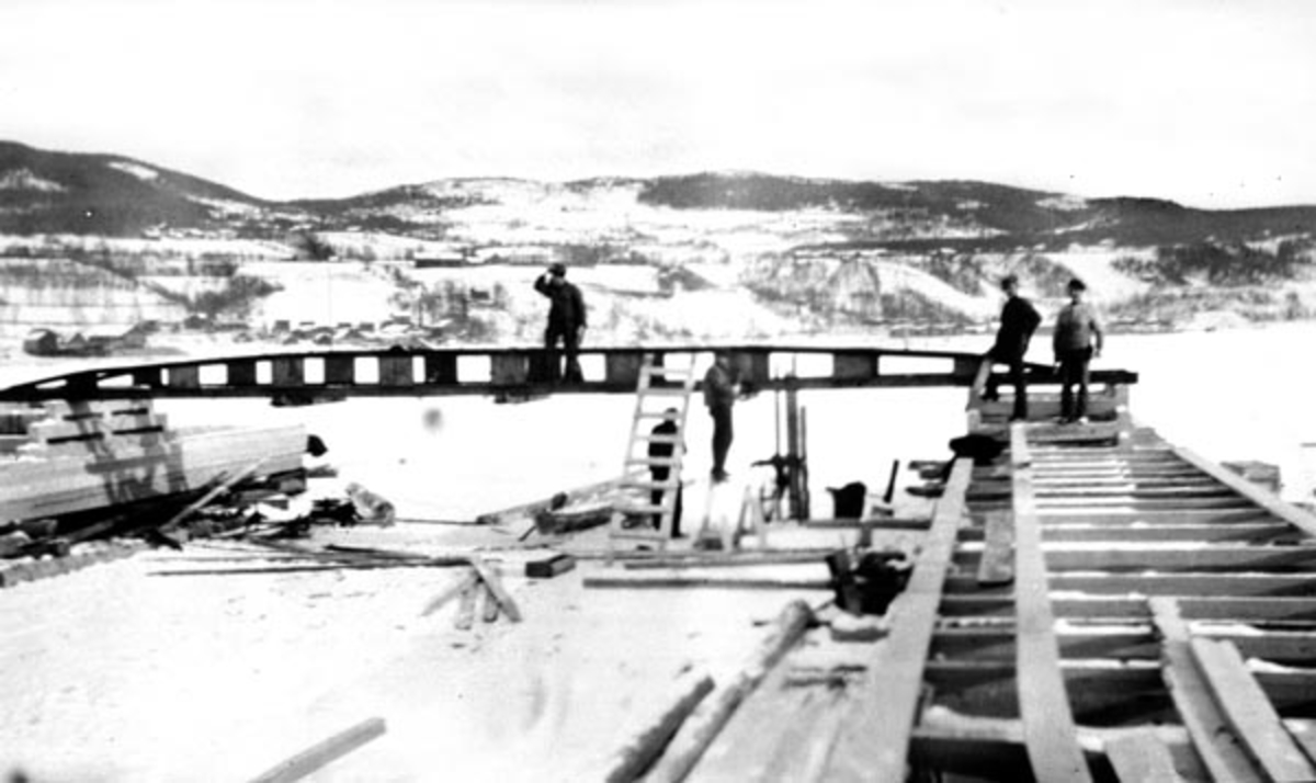 Bygging av det første soppeapparat ved Lillehammer lense, der Gudbrandsdalslågen møter Mjøsa. Dette soppeapparatet ble bygd i 1909. Det er av den typen Arnt Svartedal (1869-1909) utviklet. I slike apparater ble tømmeret ledet inn i en kanal mellom to flåteganger, med stokkenes lengderetning vinkelrett på strømretningen. De to flåtegangene var forbundet med tre stålbruer, hvorav den øverste og den ytterste sto fast, mens den midtre var bevegelig på en skinnegang langsetter de nevnte flytegangene. Bruene hadde nedfellbare bein, såkalte «sverd», og den bevegelige brua var forbundet med en motorisert vinsj. Denne kunne brukes til å presse tømmer mot den nedreste, faste brua. Dermed kunne det slås vaiere rundt tømmerbunten, før sverdene på den nederste brua ble løftet slik at de ferdige buntene eller «soppene» kunne slippes ut av moseapparatet, som da var klart for neste sopping. Dette fotografiet er tatt mens soppeapparatet var under bygging.  Vi ser rammeverket for flyegangene og den nederste, faste brua, samt en del av arbeiderne som var med og bygde innretningen. I bakgrunnen ser vi landskapet på østsida av Mjøsas nordende, det den gang landlige Søre Ål, som seinere er blitt et boligområde i Lillehammer by.

Da Kristiania Tømmerdirektion i 1903 vedtok å overta administrasjonen av fløtinga på Mjøsa hadde Lillehammer lense lenge hatt lokalt eierskap. Den nevnte beslutningen fikk i første omgang ingen annen konsekvens for lensedriften enn at det fra da av var Tømmerdirektionen som inviterte til anbud på driften av de 8 lenseanleggene som fantes der fløtingsvassdragene rant inn i innsjøen. I 1908 inngikk Kristiania Tømmerdireksjon forpaktningsavtale for Lillehammer lense, og det var denne organisasjonen som moderniserte anlegget. De to første sesongene, i 1909 og 1910, ble soppeapparatene drevet av en dampmaskin. Etter dette inngikk Tømmerdireksjonen avtale med Lillehammer kommunale elektricitetsverk om leveranser av elektrisk energi. Fra da av ble soppinga av tømmer utført med elektromotorer som drivkraft. Trepongtongene vi ser på dette fotografiet ble erstattet av stålpongtonger fra Hamar Jernstøperi og mek. Verksted våren 1916.

Onsdag 12. mai 1909 beskrev avisa «Lillehammer Tilskuer» det nye soppeapparatet ved byens lenseanlegg slik:

«Soppemaskinen ved Lændsen er nu paa det nærmeste færdig. Det bestaar av to 64 Meter lange flydende Broer, der holdes sammen av to 12 M. lange Tverbroer. Paa den ene Langside ligger Maskinen. Hele Apparatet ligger paa langs i Strømretningen. Tømmeret kommer drivende ind i den øvre Ende, og føres af Strømmen ned mod to flere Meter dybe Arme, der stikker ned fra den nederste Tverbro. Her hobes det da op, og det slig, at Stokkene blir liggende paa tvers imellem begge Langbroer. Naar hele Rummet er fyldt trækkes en forskyvelig Bro, der ogsaa er forsynet med nedstaaende Arme, nedover af Maskinen, og derved presses Tømmeret sammen i en flere Meter dyb Sop. Der slaaes Staaltraadbaand om, og Soppen er færdig. Armene paa den nederste Tverbro løftes op og Soppen flyder ud. Saa begyndes der paa igjen paa samme Maade.»