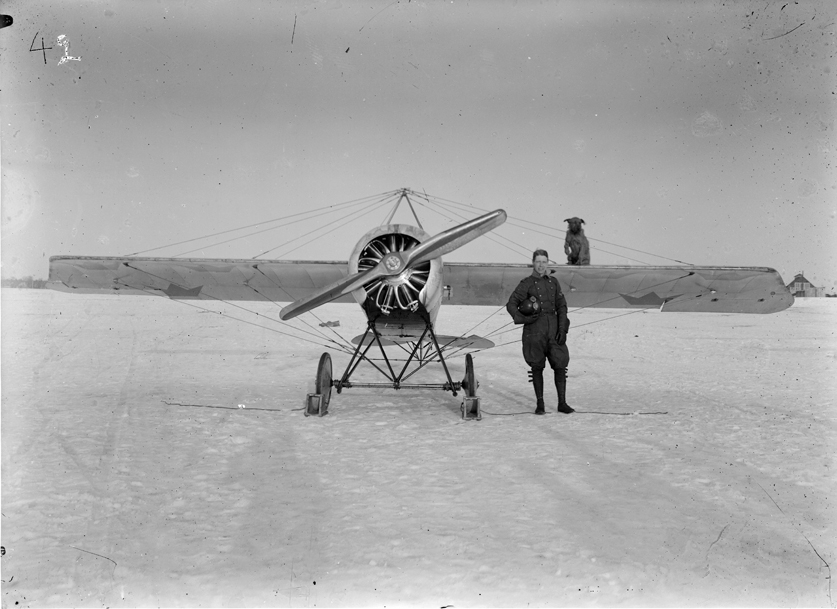 Flygplan Thulin K "Thulinjagaren" på marken. En man står framför flygplanet och en hund sitter på flygplansvingen.
