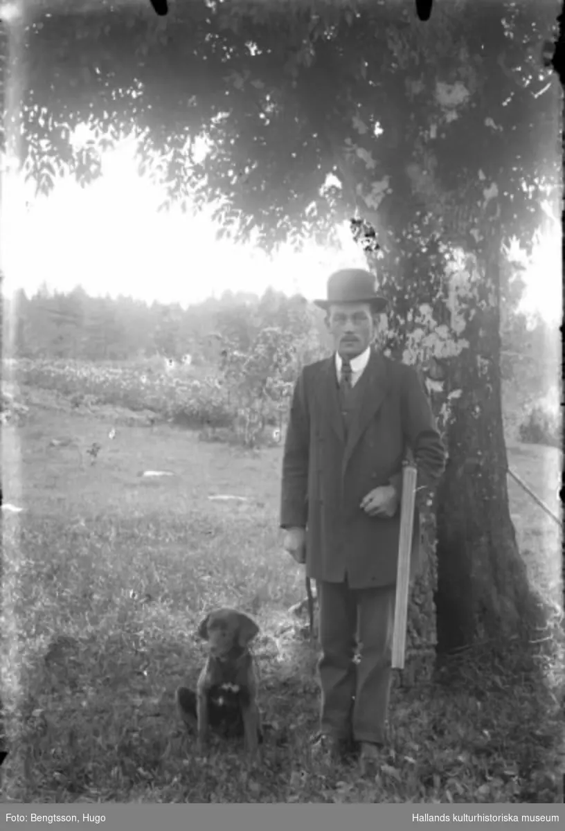 Karl Nilsson står finklädd i hatt med hund och gevär vid ett träd.