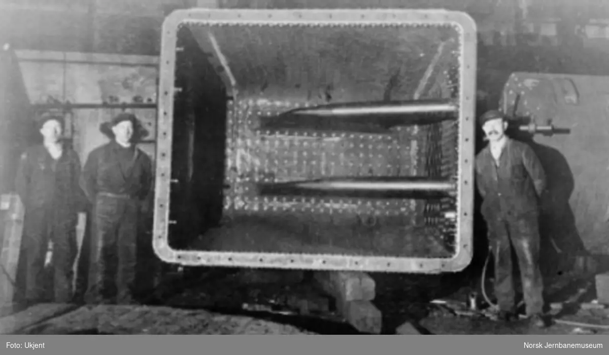 Damplokomotiv type 49a "Dovregubben" under bygging; tre arbeidere poserer med fyrkasse med syphoner (vannsirkulasjonsrør)