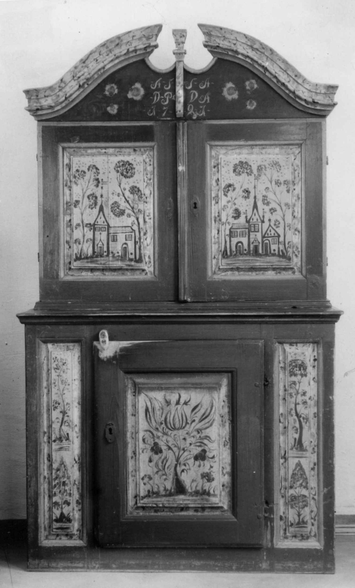 Framskap i to deler. Buet topplist. Skuffer og skjehylle i overskap. Malt innvendig og utvendig. Kirke og bygning malt på dør på overskap. Planter på underdel. Marmorert på lister på overskap.