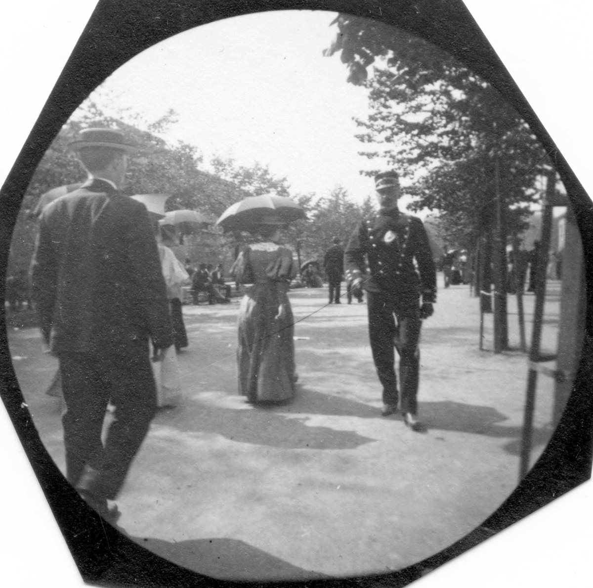 Mennesker spaserer langs Karl Johans gate, Oslo.  Mann med dress med ryggen til, damer med parasoller og rittmester Michelet i uniform kommer mot fotografen.