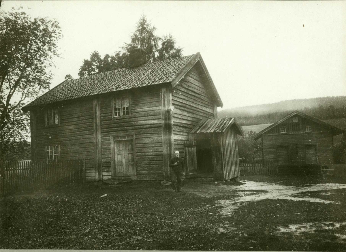 Åserud (Tømte), Eidsvoll, Øvre Romerike, Akershus. Mann spaserer over gårdsplass foran våningshus i tømmer. Stabbur i bakgrunnen.