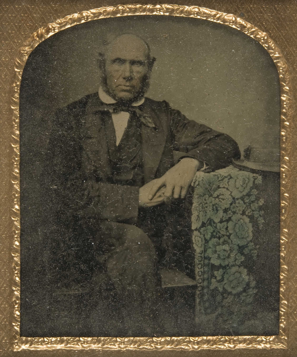 Portrett av mann, skipsreder og skipsfører Mads Høegh Johnsen (1806-1891), fra Slagen, Tønsberg, Vestfold. Han sitter med venstre arm hvilende på bord med duk og hatt, knebilde. Duken og hudpartier antatt kolorert.