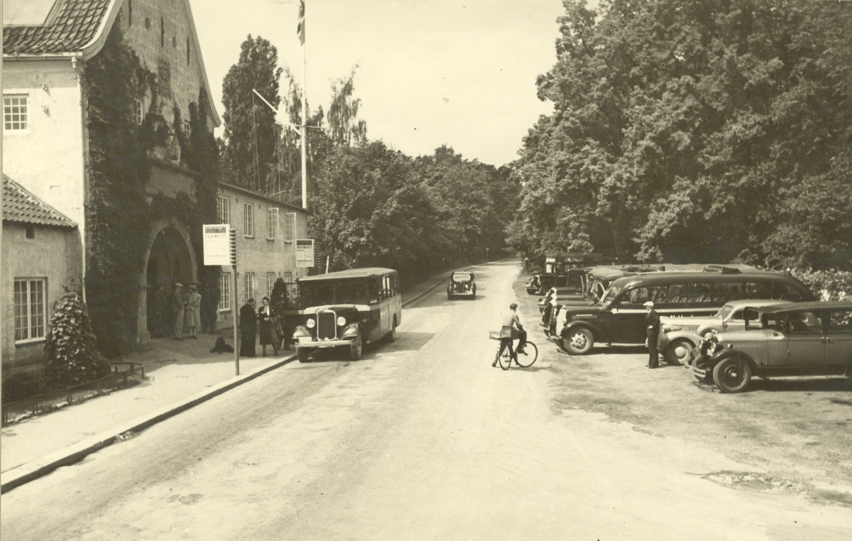 Hovedinngang med stadsporten etc. sett fra Museumsveien. Biler og lave busser står på parkeringsplassen. Norsk Folkemuseum, Oslo 1939.
