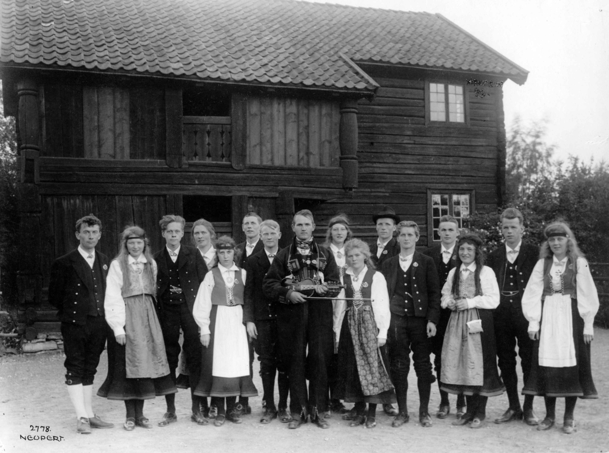 Folkedansstevne 1925. Dansere i folkedrakter. Felespiller i midten. I bakgrunnen tømmerhus.