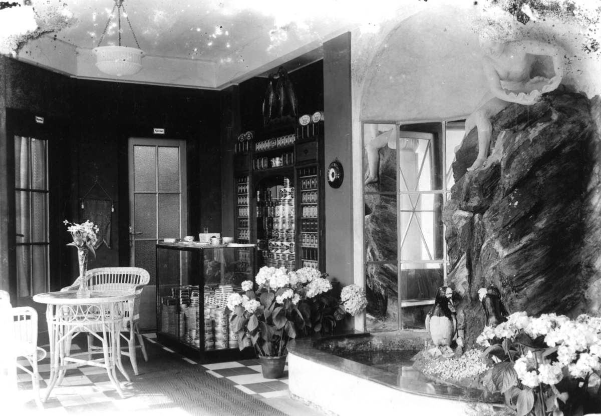 Jubileumsutstillingen på Frogner, Oslo, 1914. Interiør paviljonger. Skulptur til høyre i bildet. Blomster. Reklameprodukter i hyller i bakgrunnen.
