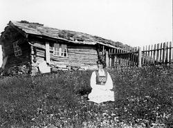 Kvinne i folkedrakt sittende utenfor våningshus, ukjent sted