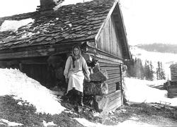 Pike/kvinne (budeie?) med geit sittende utenfor uthus, antat