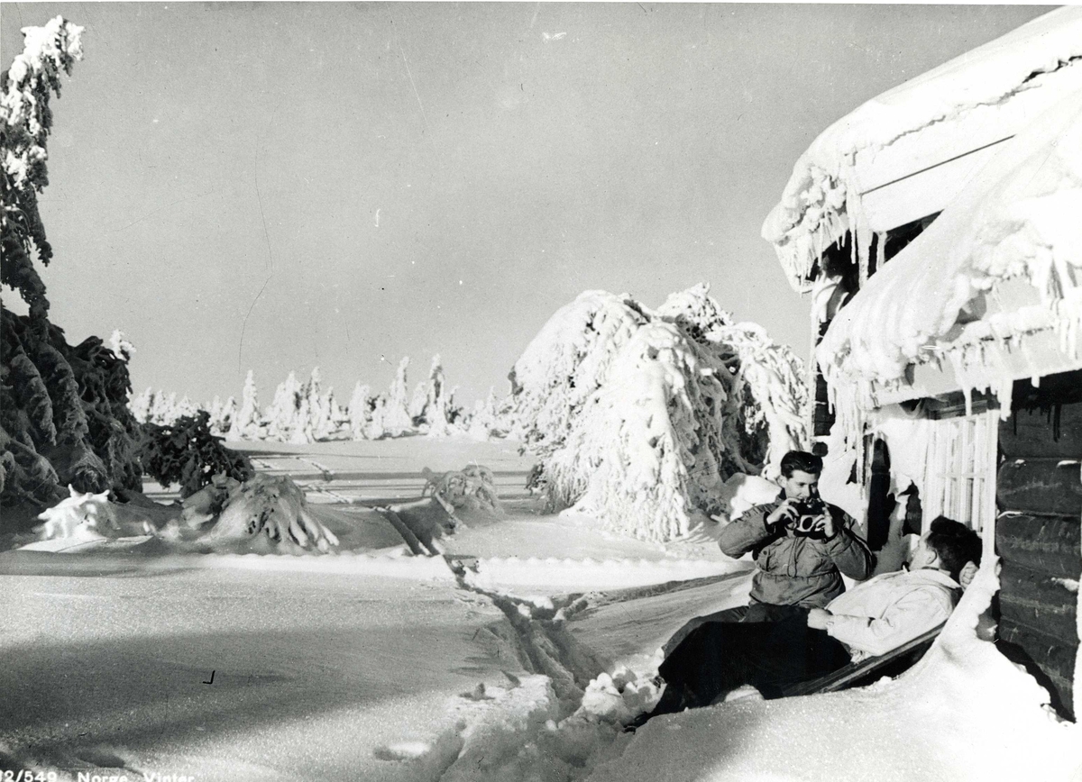 Avfotografert postkort. To skiløpere i hytteveggen i snødekt skogslandskap.  En har et fotoapparat.