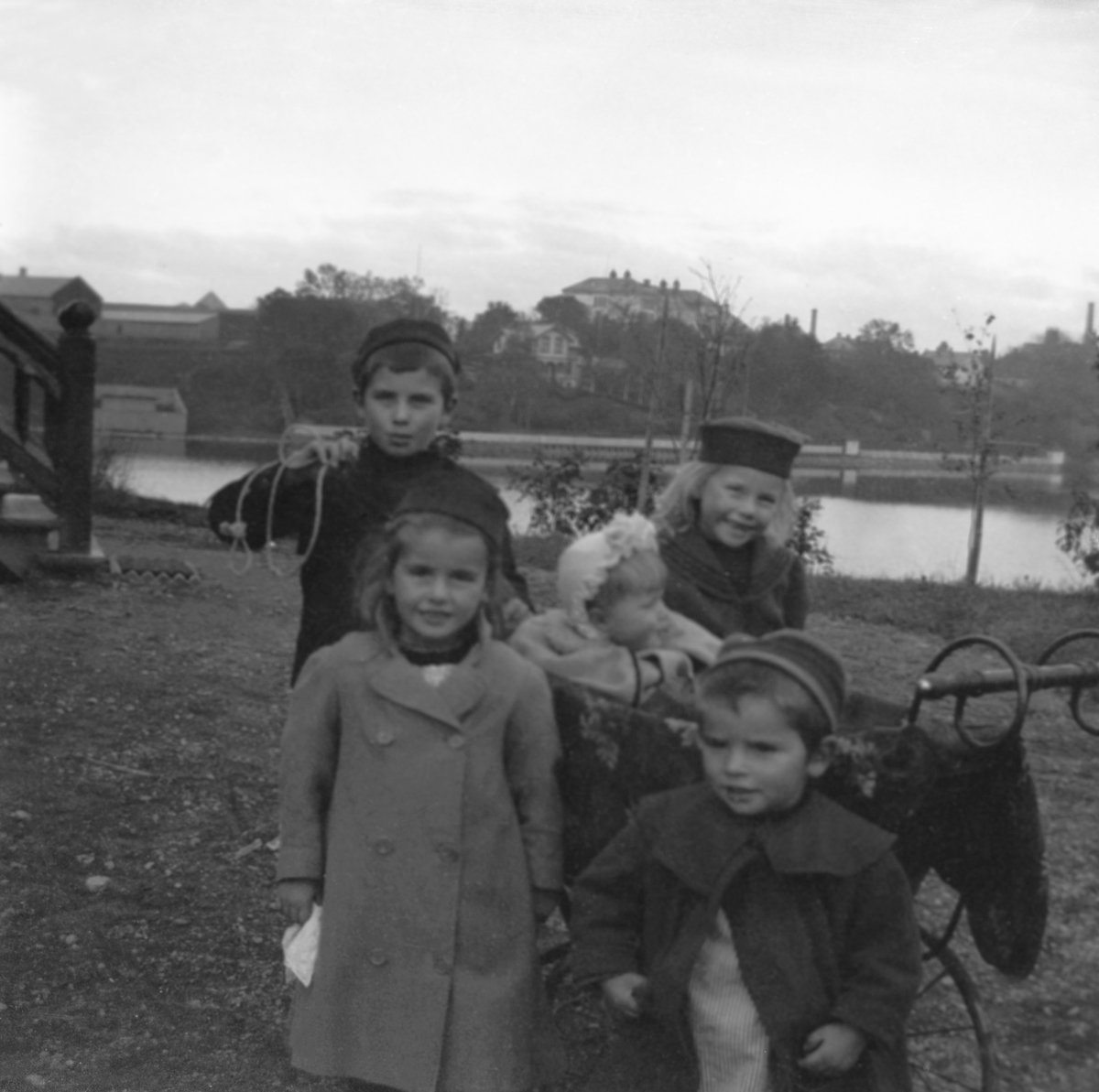 Fritjof, Guri, Nanna i barnevogn, Sofie og Thorleif Arentz ved kjøkkeninngangen til hjemmet Elverhøy i Trondheim. Nidelven i bakgrunnen. Fotografert 1902.