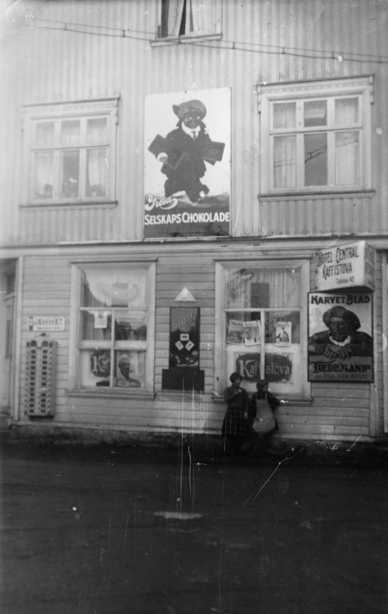 To jenter utenfor Lyngdal Kaffestove og Hotel Central i Lyngdal 1934. Reklameskilter fra Freia og Tiedemann på veggen.
