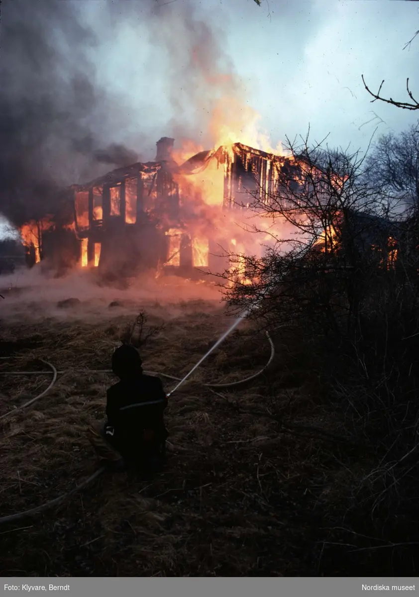 Ett brinnande hus på Lidingö. I förgrunden en brandman med vattenslang.