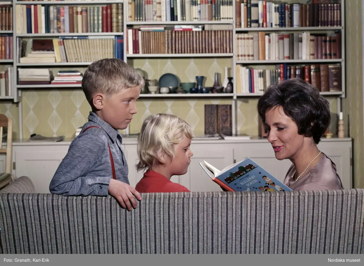 Vardagsrum. En kvinna och två barn sitter i soffa framför bokhylla och läser ur "Bullerbyboken" av Astrid Lindgren