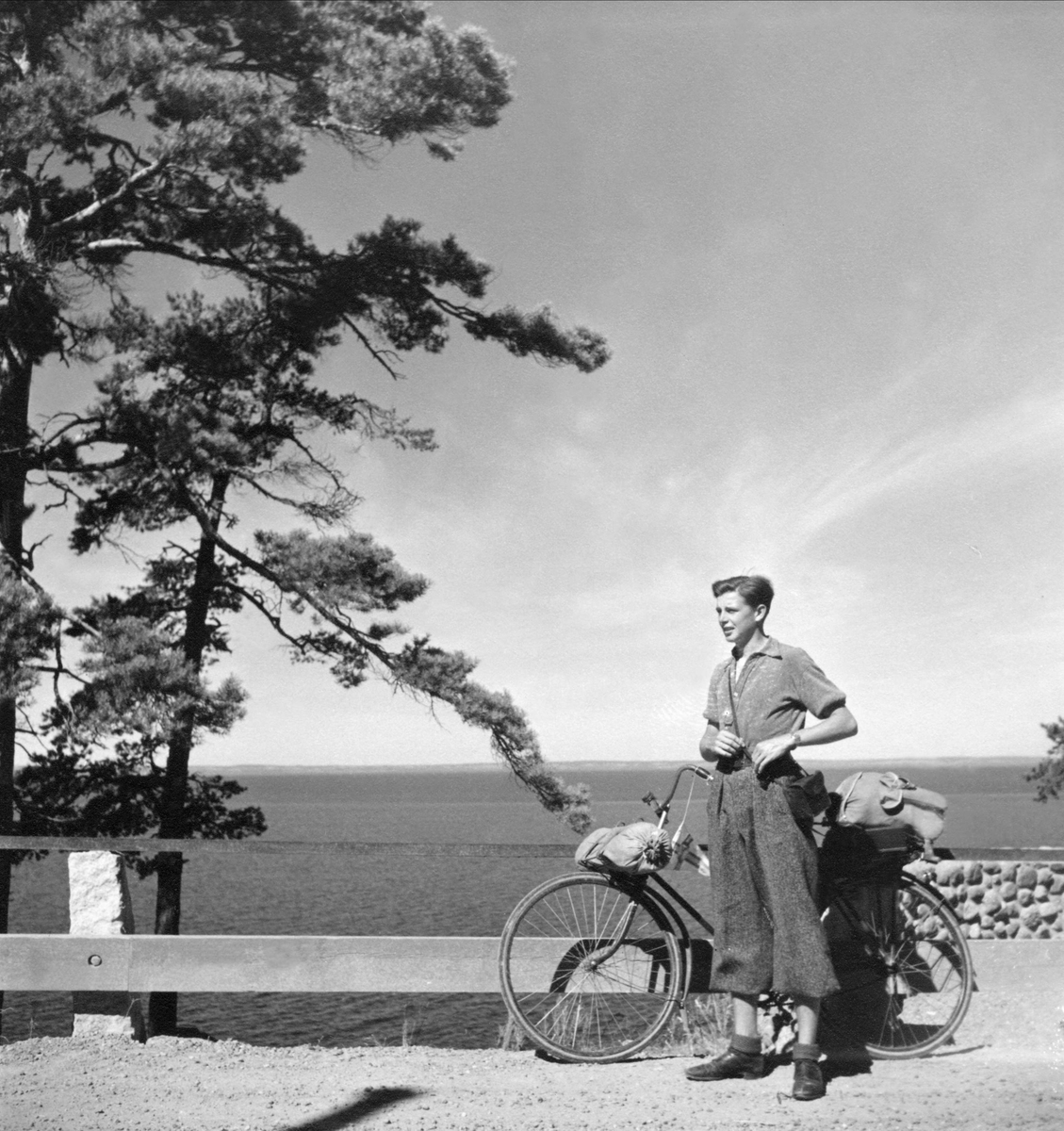 Cyklist, ung man, i skjorta och knäbyxor som stannat vid en utsiktsplats intill en sjö.