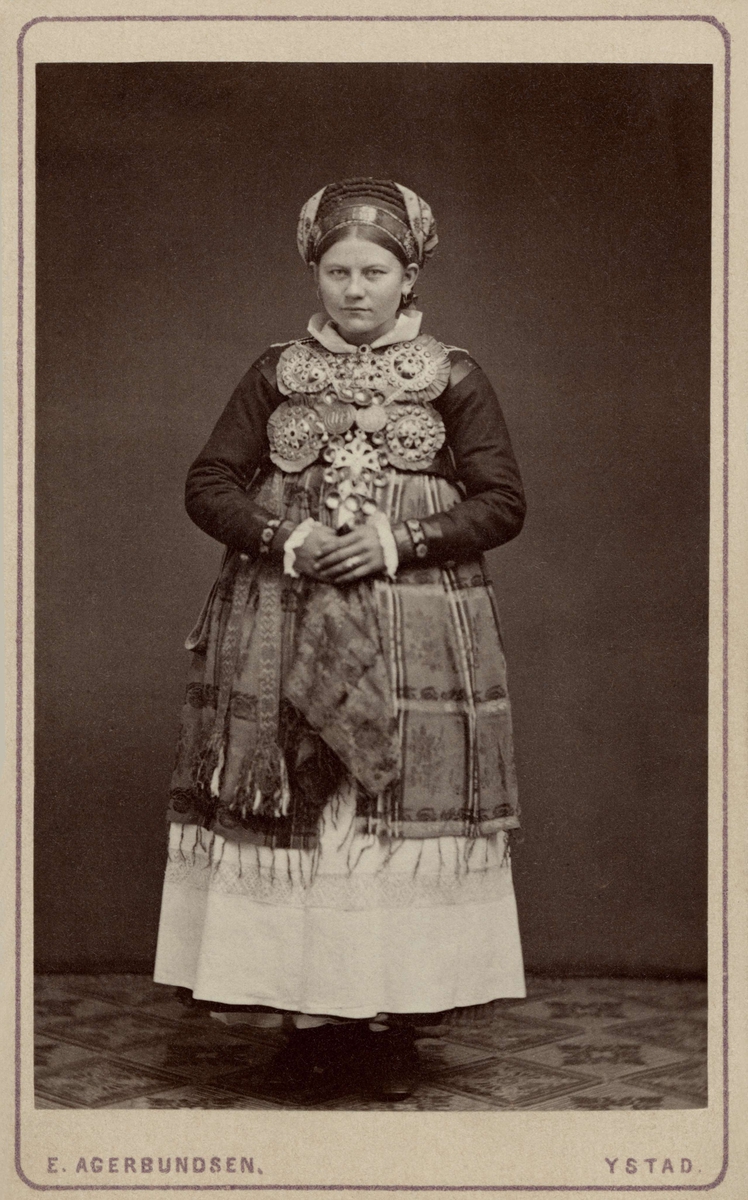Porträtt av stående kvinna iklädd folkdräkt. På kortet är noterat att hon bär bruddräkt från Ingelstads härad i Skåne. Nordiska museet inv.nr. 18541C