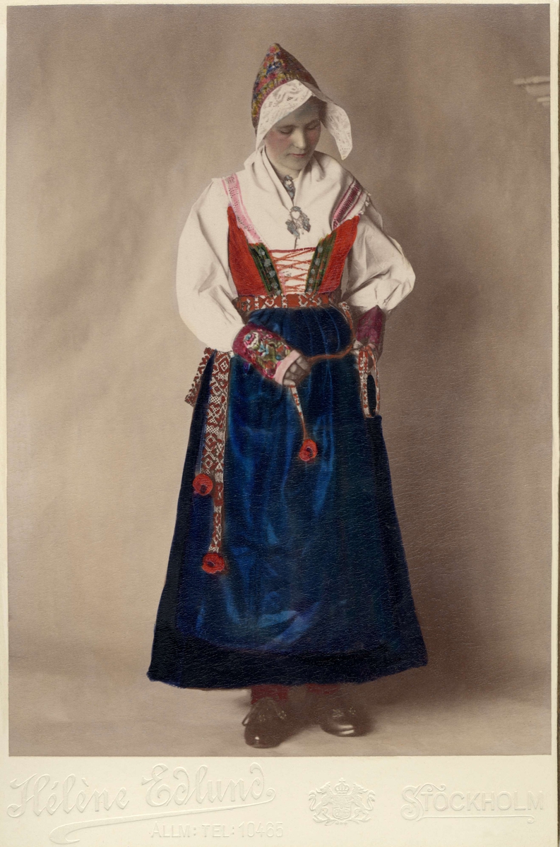 Porträtt av kvinna klädd i Åls högtids- och kyrkodräkt, Dalarna, Nedan Siljans fögderi. Handkolorerat fotografi.
