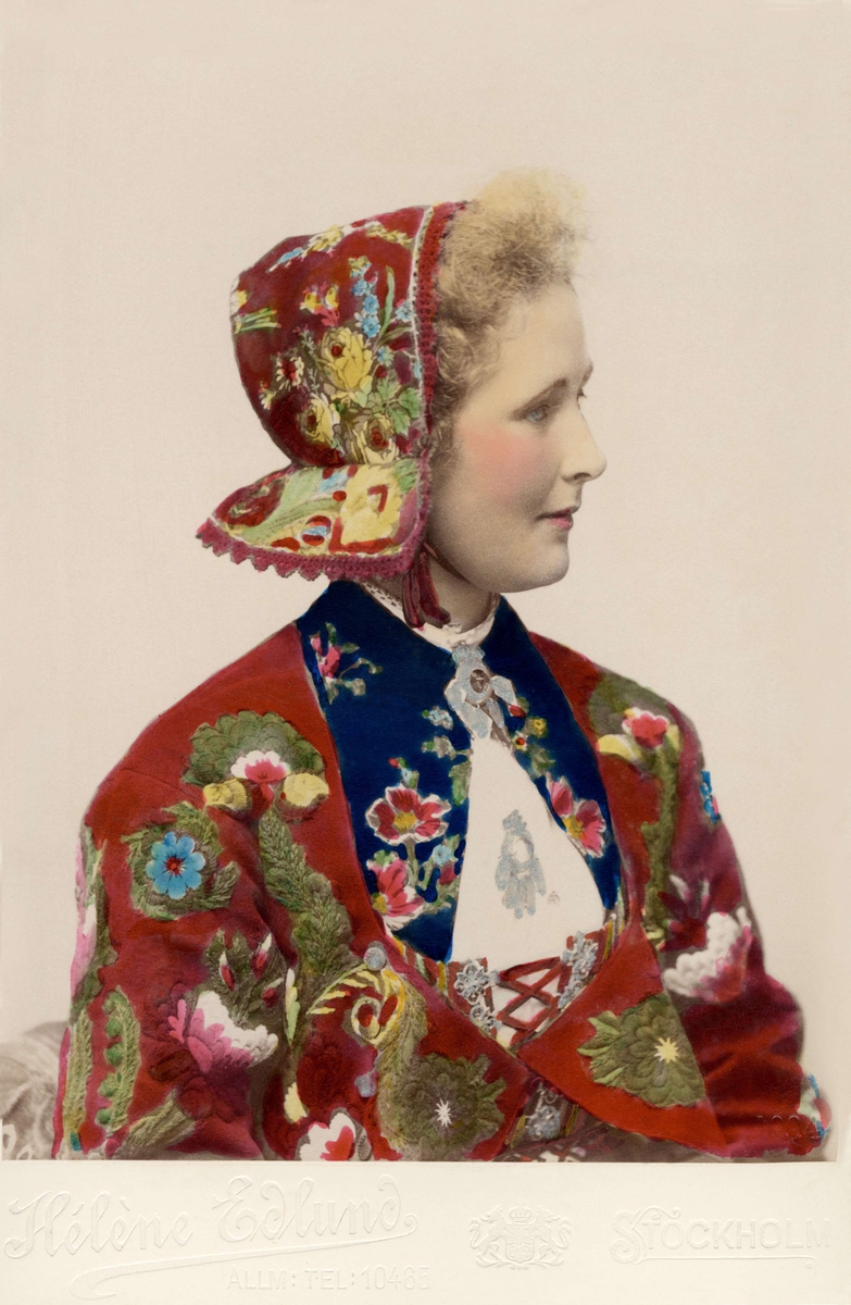 Porträtt på en kvinna i blommig jacka och hätta. Klädd i dräkt från Dala-Floda, Västerdalarna med broderad s.k. påsömströja och dito hätta.