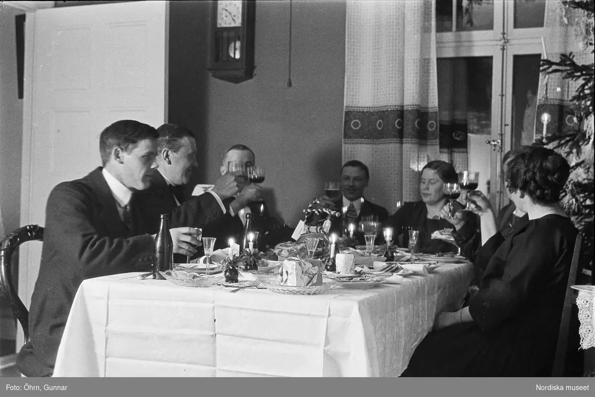 Julafton hos familjen Karlberg i Stockholm. Sällskap som sitter vid  matbord höjer sina glas till en skål. Fotografen själv längst till vänster i bild.