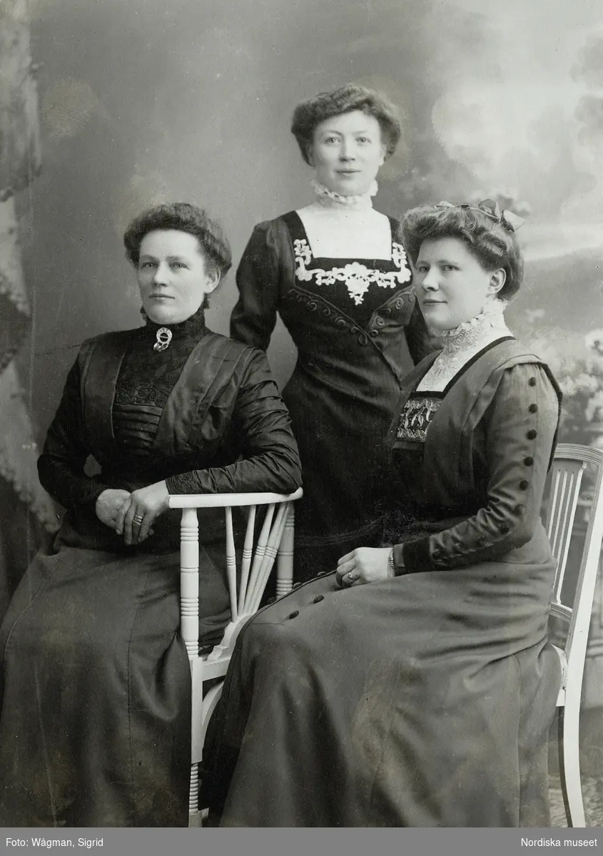 Ateljéporträtt av tre medelålders kvinnor i klänningar med broderier och höga spetskragar. Kabinettkort.