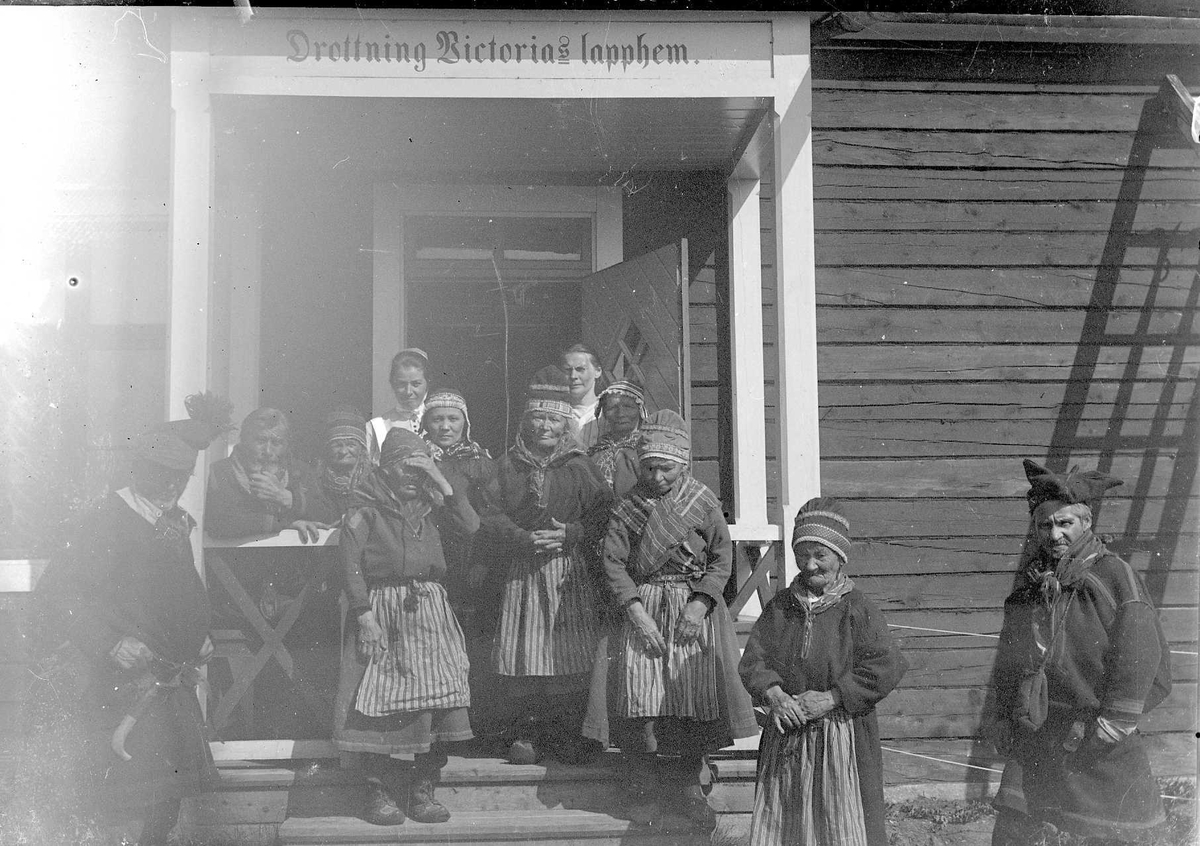 Gruppbild på yttertrappan till det så kallade "Drottning Victorias lapphem". Äldre samer vid drottning Victorias sjukhärbärge och ålderdomshem i Karesuando, dörröppningen i bakgrunden står två sköterskor.