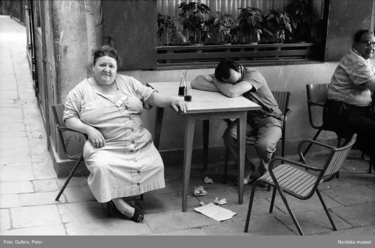 Kvinna och man sitter vid kafébord. Vin i glas och karaff på bordet. Mannen sover med huvudet i armarna. Kvinnan ser in i kameran. Neapel, Italien