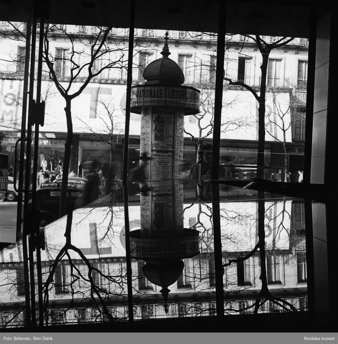 Utsikt mot gatan och en reklampelare genom ett skyltfönster i stadsmiljö. Speglingar.