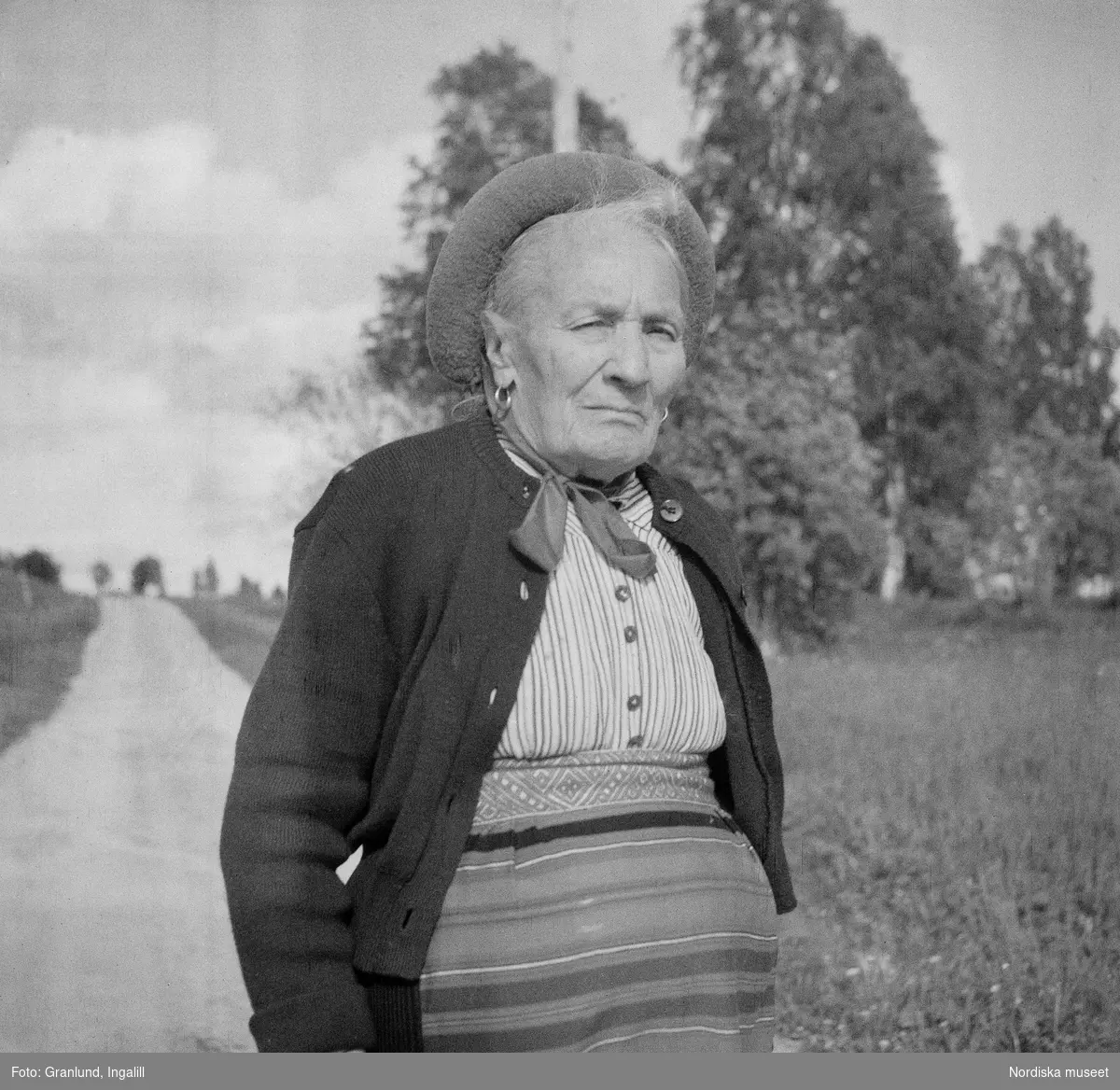 Pergården (tidigare Sjonsgård) i Osmundsbergs by i Boda socken, Dalarna. Fräs Kerstin (Kirstin) Jonsson klädd i yllekofta och smårandig blus, men med mössa, kjol och förkläde enligt Boda-sed.