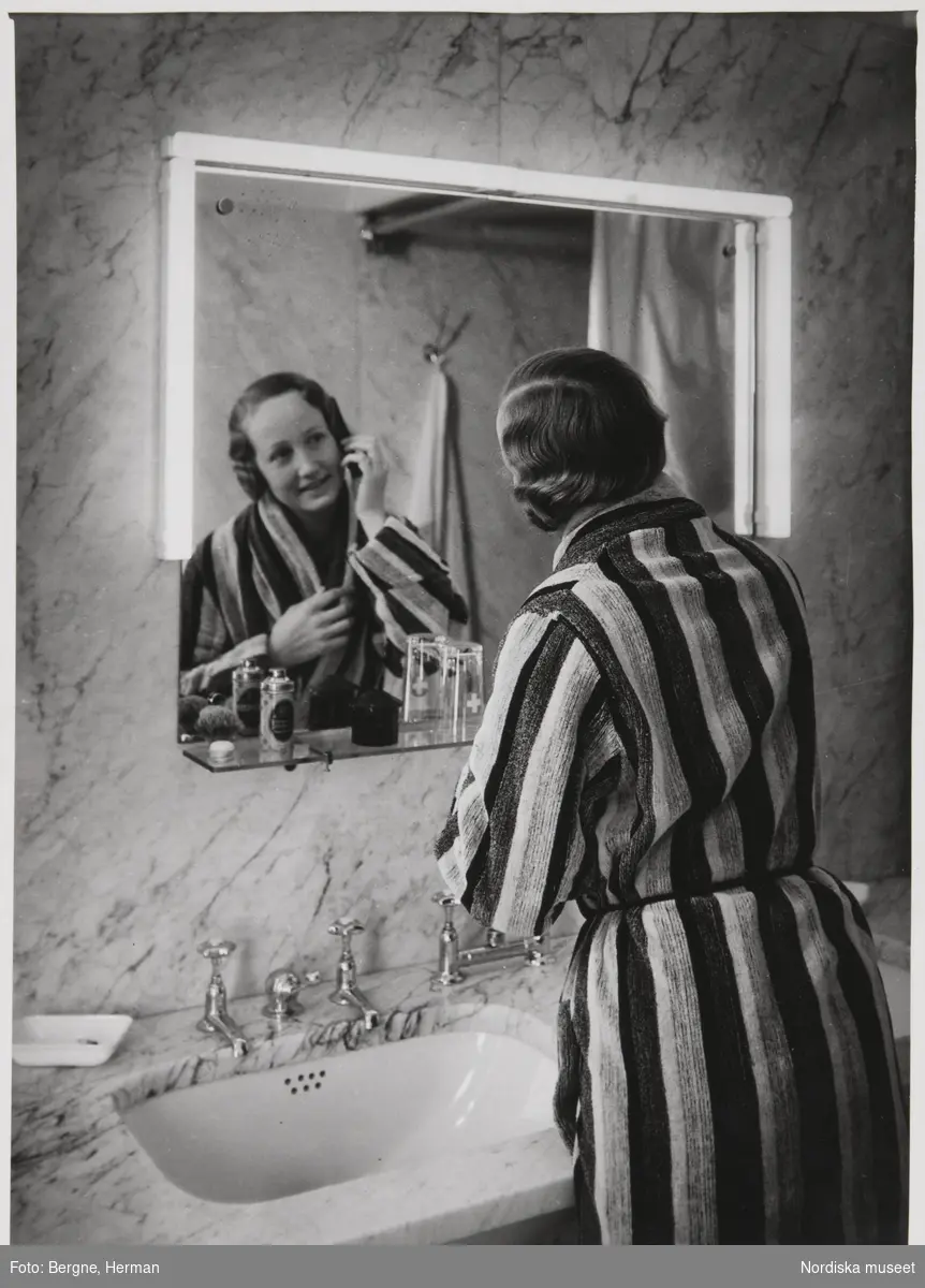 Interiör från badrum. Kvinna i randig morgonrock med handen vid sitt högra öra. Ljuset vid spegeln 1935, s. 4.