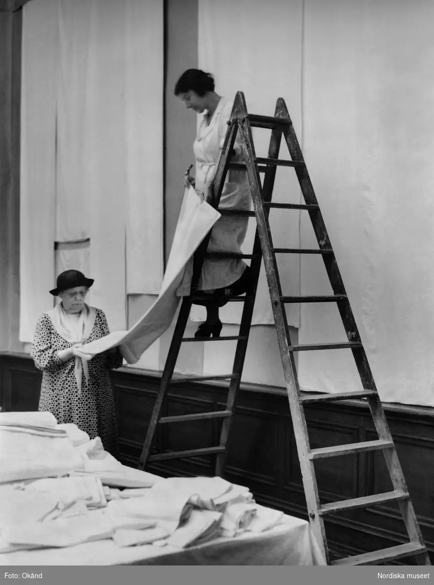 Elisabeth Thorman till vänster under arbetet med damastutställningen på Konstakademien i Stockholm 1936. Gunhild Thörn på stegen hjälper till med hängningen av alla inlånade dukar.