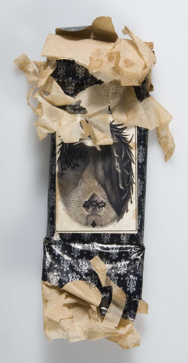 Begravningskaramell med omslag av beige silkespapper och svart glanspapper med tryckta silverfärgade medaljonger. Pappersetikett med tryck symboliserande treenigheten.