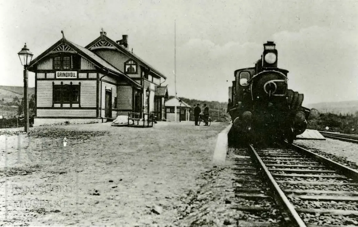 Grindvoll jernbanestasjon. Stasjonsbygningen til venstre, jernbanesporet med et lokomotiv til høyre. To menn står mellom toget og huset. 