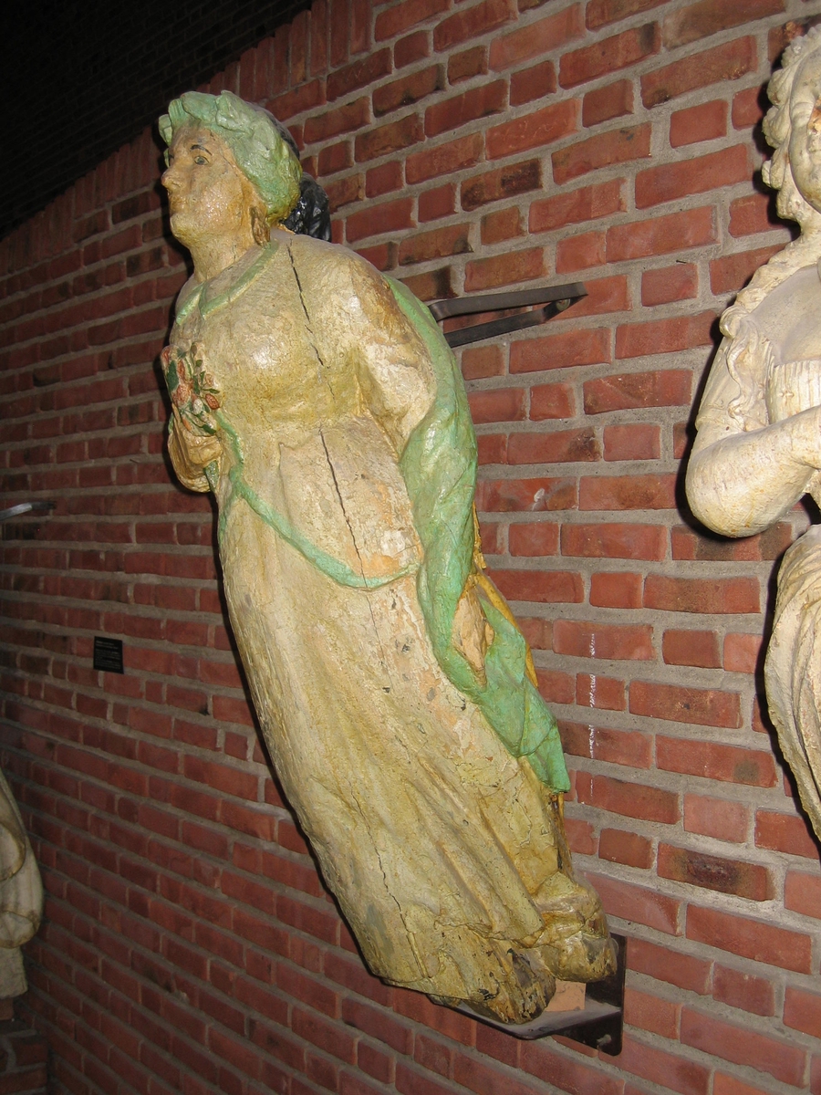 En klassisk gallionsfigur, venstre arm ned langs siden, høyre arm bøyd med en blomsterbukett i hånden mot brystet. Sort hår, hvit kjole med grønn kant, blomster i høyre hånd mot brystet.
