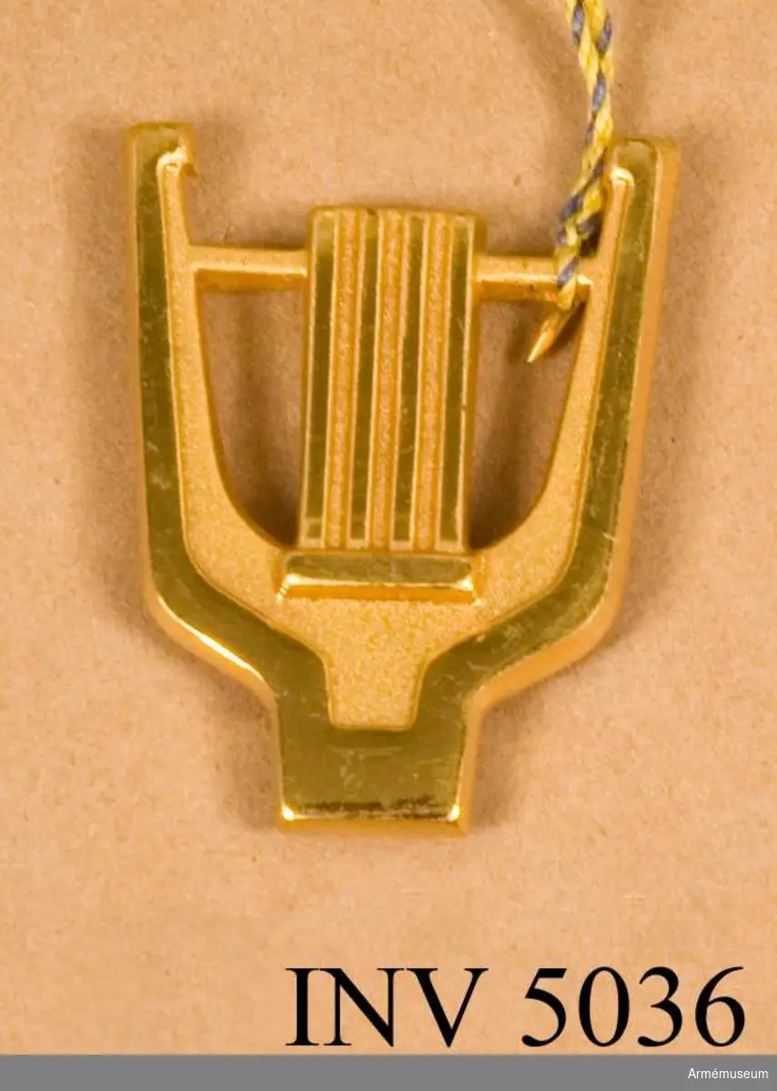 Tillverkat av metall och i metallfärg m/1960 guld. Utföres i två storlekar. Lyran som symbol för musikpersonal har behållits men här har ädelsmeden Sigurd Persson gjort en helt ny utformning. 
