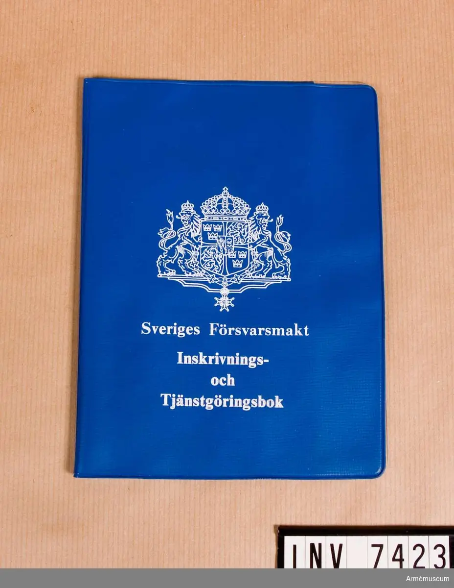 En pärm i blå plast med på framsidan i vitt tryck: Det stora riksvapnet, Sveriges Försvarsmakt Inskrivnings- och tjänstgöringsbok. På insidan fack för boken mm. 
