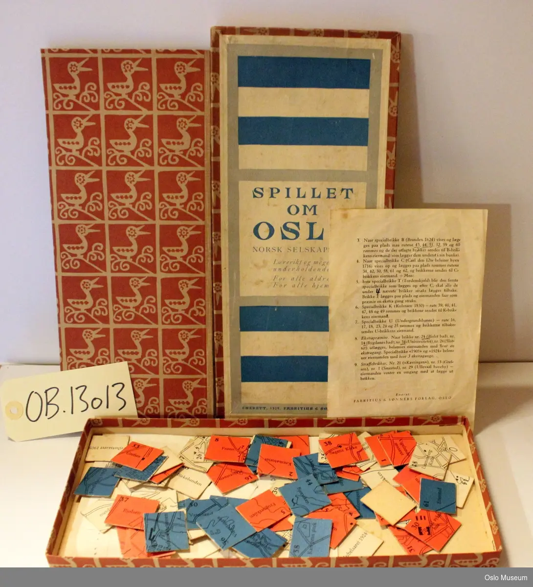 Rektangulært brettspill i eske med 72 brikker og spilleregler. Brun eske, "Spillet om Oslo" i blått på hvit bunn.