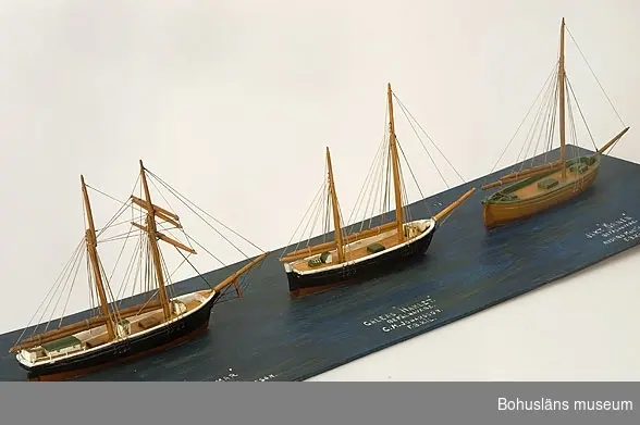 Tre fartygsmodeller, skonerten Hjalmar, galeasen Hamlet och jakten Örnen från Fiskebäckskil.
Tillverkade av kapten John Emil Olsson (1880-1950), Fiskebäckskil på Skaftö i Lysekils kommun på 1940-talet.
Trä, metall, lintråd.
Jakten - höjd 20 cm, längd 24 cm, bredd 4,5 cm; platta 79 x 22 cm; okänd skala.
Vattenlinjemodeller med stående och löpande rigg, däckshus och lastluckor.  Skonerten och galeasen med svartmålade skrov och röd bottenmålning, jakten med gulmålat skrov och röd bottenmålning. Monterade på utskuren blåmålad platta.
Ovanpå  plattan står textat med vita tryckbokstäver från vänster till höger: 
SKONERT "HJALMAR" BEFÄLHAVARE. SJÖKAPTEN. ANTON KRISTENSSON. F.B.KIL
GALEAS "HAMLET" BEFÄLHAVARE. C.M. JOHANSSON. F.B.KIL.
JAKT "ÖRNEN" BEFÄLHAVARE ANDERS MARTINSSON. F.B.KIL.

Definitioner:
Skonert
Ett segelfartyg med två eller fler master av vilka den förligaste har fullständig råsegeltackling och övriga gaffelsegel med trekant-toppsegel.
Råsegel - rektangulärt eller trapetsformigt segel som förs på en rå, dvs. en horisontell stång fäst vid mastens framkant
Gaffelsegel - trapetsformat segel fäst längs akterkanten av masten och med sina övre (kortare) och undre (längre) lik (kanter) fäst vid två rundhult (gaffel resp. bom). 
Galeas
Ett mindre, tvåmastat segelfartyg riggat med gaffelsegel som skonare med stormasten främst. 
Gaffelsegel - trapetsformat segel fäst längs akterkanten av masten och med sina övre (kortare) och undre (längre) lik (kanter) fäst vid två rundhult (gaffel resp. bom). 
Jakt
Enmastat segelfartyg med öppet däck och en mindre kajuta akterut. 
Gaffelsegel - trapetsformat segel fäst längs akterkanten av masten och med sina övre (kortare) och undre (längre) lik (kanter) fäst vid två rundhult (gaffel resp. bom).

Ur handskrivna katalogen 1957-1958:
"Hjalmar", "Galeas", "Hamlet"
Skonerten Hjalmar, Galeasen Hamlet, Jakten Örnen. Modell på platta. Plattans mått 78,5 x 22,5. Föremålen hela.
Från kapten Olssons saml., Fiskebäckskil.

För ytterligare information om John Emil Olsson och förvärvet, se UM005807.

Föremålet presenterat på Bohusläns museums hemsida år 2009 - 2013, webbutställning "John Emil Olsson Fiskebäckskils sjöfartsmuseum".