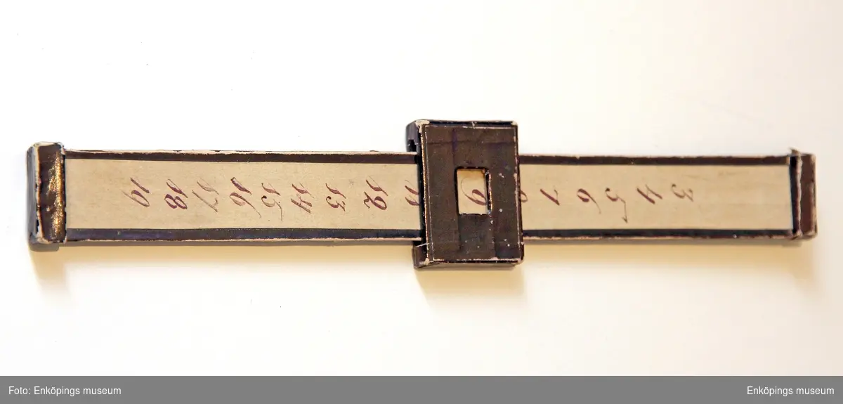 En svart markeringstång klädd i vaxat  papper med siffrorna från 3- 19 skrivna med tusch. Runt stången finns det en rörlig fyrkantig öppning i mitten.