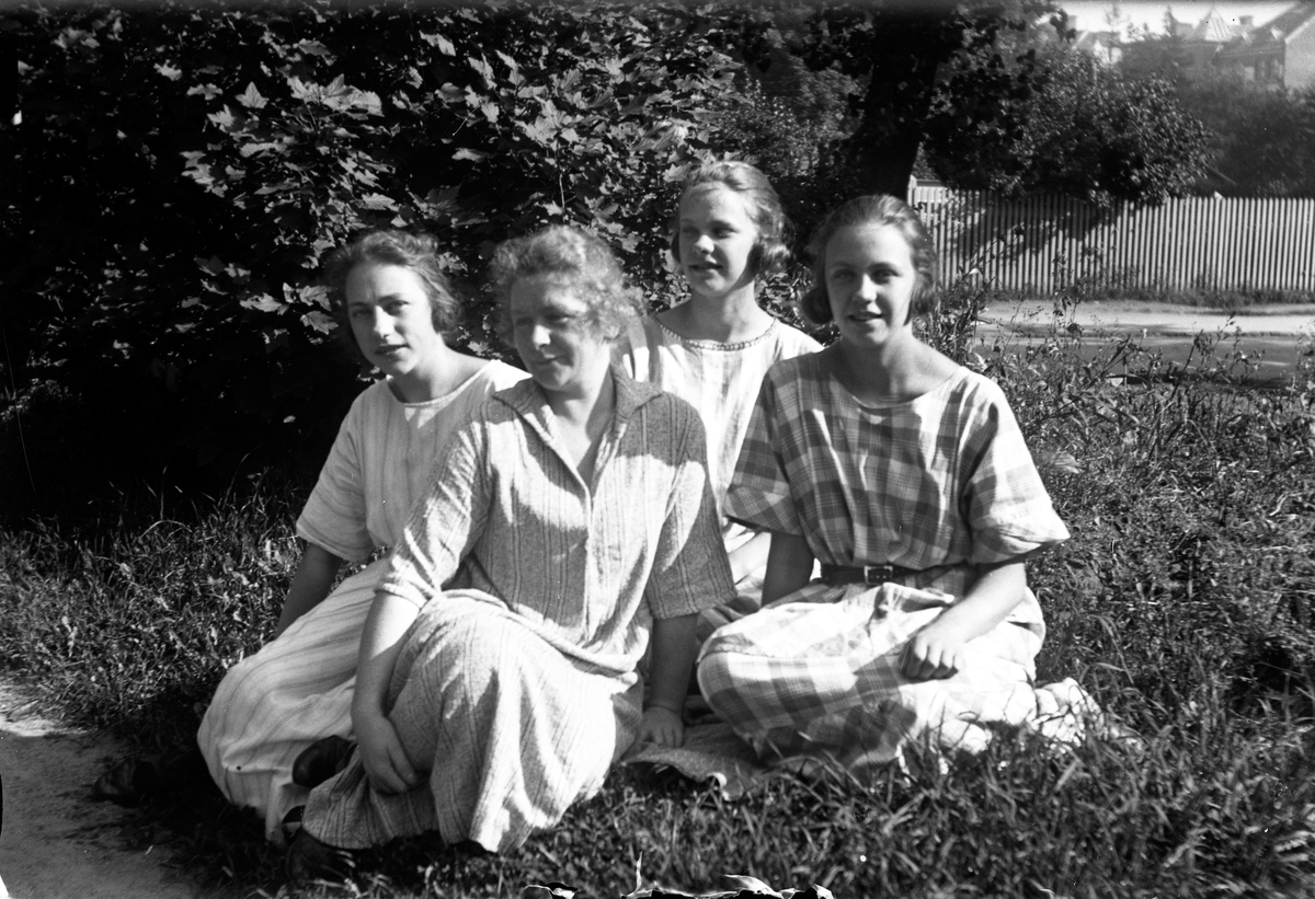 Fyra kvinnor sitter i gräset vid ett träd, i bakgrunden syns hus och ett staket.